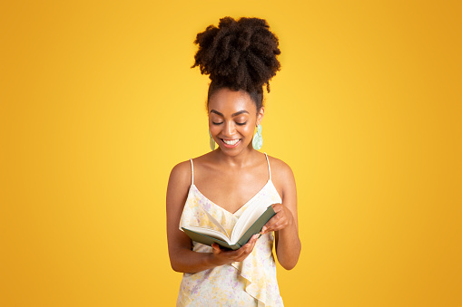 Smiling millennial african american lady reading book, enjoy free time, homework, isolated on yellow background, studio. Ad and offer, study, hobbies, education and day planning