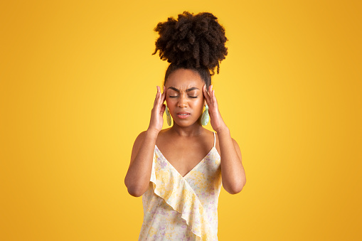 Sad tired millennial african american lady puts hands to temples, suffering from headache, isolated on yellow background, studio. Migraine, health problems, overwork and stress