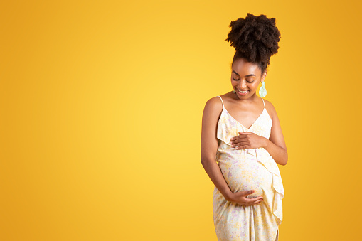 Glad young black woman touching big belly, feels baby move, enjoy pregnancy, isolated on yellow background, studio. Lifestyle, sale, ad and offer, expectation of child, family planning