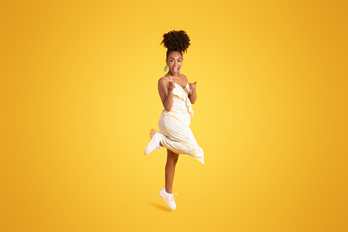Excited cheerful young african american lady in dress jumping, has fun, point fingers to camera, isolated on yellow studio background. Your choice, turn, lifestyle and fashion, sale, ad and offer