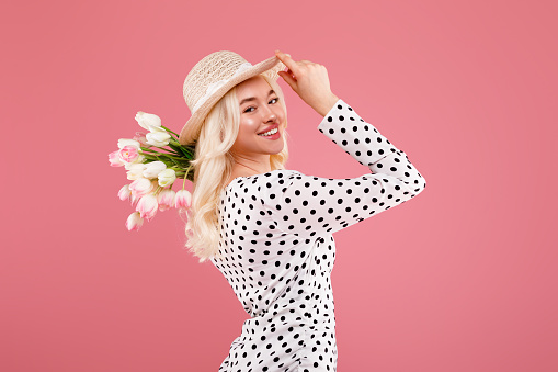 Beautiful young lady wearing dress and straw hat, posing with flower bouquet and smiling at camera, standing isolated over pink background