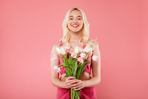 Happy charming blonde lady holding bouquet of flowers, enjoy women day or birthday, smiling at camera, posing on pink studio background. Human emotions, congratulations