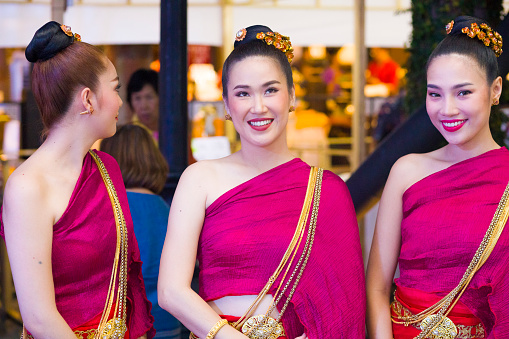 Capture of three traditionally dressed and smiling thai women. They  are standing in public to show fashion and elegance style. Two are toothy smiling and looking. One is looking back. Scene is at One NImman Square in Chiang Mai. Women are dressed in elegant red. Hair of all is made in same style. Women are part of cultural entertainment on night market to visitors and tourists. Woman are standing for being photographed by people and anyone while demonstrating thai culture and fashion. In background are people and visitors of night market