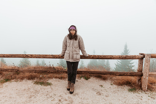 A young female hiker is walking in nature and searching for a path on her mobile phone map. She is in a forest where it's foggy, cold, and windy. She leans against a wooden mountain fence.