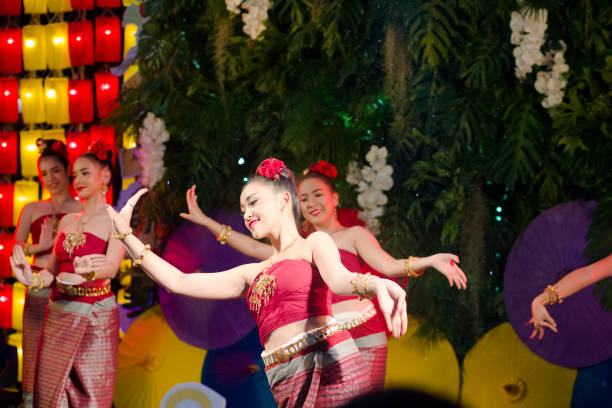 Rum Thai dancing young thai women moving in slow poses  and  performance. Rum Thai dancing young thai women moving in slow poses  and  performance. Scene is on night market at One Nimman square in Chiang Mai. Women are dressed  traditional clothes and with flowers in hair. 
 Women are red dressed and are dancing classical Rum Thai. Performance is on a small stage and they are dancing in public for people who are visiting market as part of several entertainment of dancers and singers true thailand classic stock pictures, royalty-free photos & images