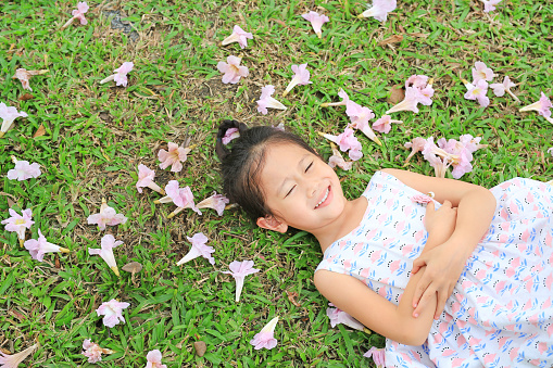 Smiling little girl lying on green grass with fall pink flower in the garden outdoor.