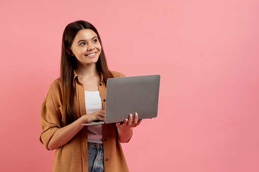 Beautiful smiling teen girl holding laptop and looking aside at copy space, young female student using computer for online education and distance learning, standing on pink background, copy space