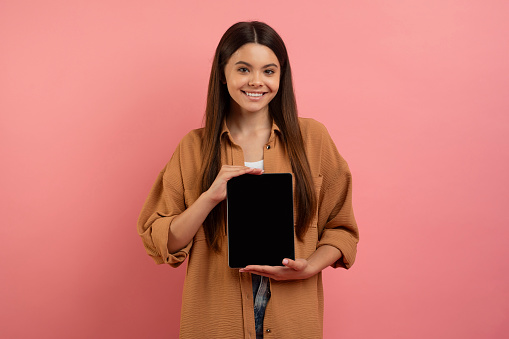 Educational App. Beautiful Happy Teen Girl Showing Digital Tablet With Black Blank Screen At Camera, Cheerful Female Teenager Recommending New Appication, Standing On Pink Background, Mockup