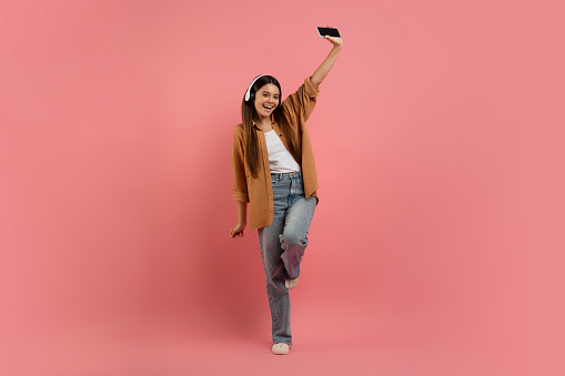 Energetic teen girl listening music on smartphone and dancing, happy female teenager wearing wireless headphones enjoying favorite song, having fun on pink studio background, full length, copy space