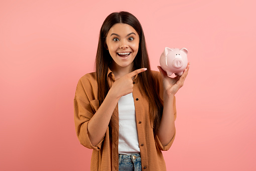 Happy excited teen girl pointing at piggy bank and looking at camera, cheerful beautiful female teenager enjoying money saving for future, standing over pink studio background, copy space
