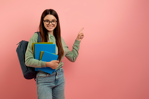 Check This. Happy Teenage Girl Holding Workbooks And Pointing Aside At Copy Space, Smiling Female Teen With Backpack And Eyeglasses Demonstrating Free Place For Advertisement On Pink Background