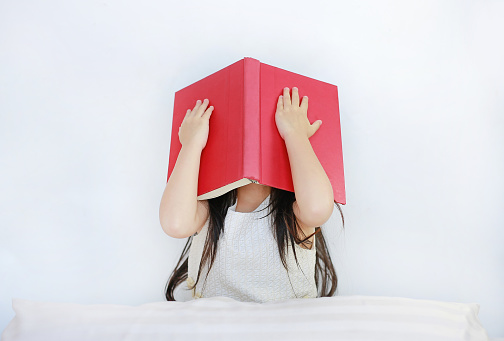 Little Asian child girl hiding face with open hardcover book sitting on bed with pillow.