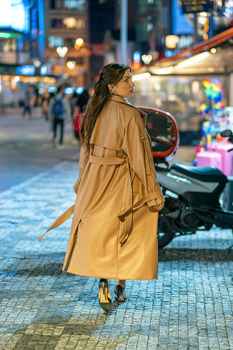 Young woman in trench coat walking on crowded street at night