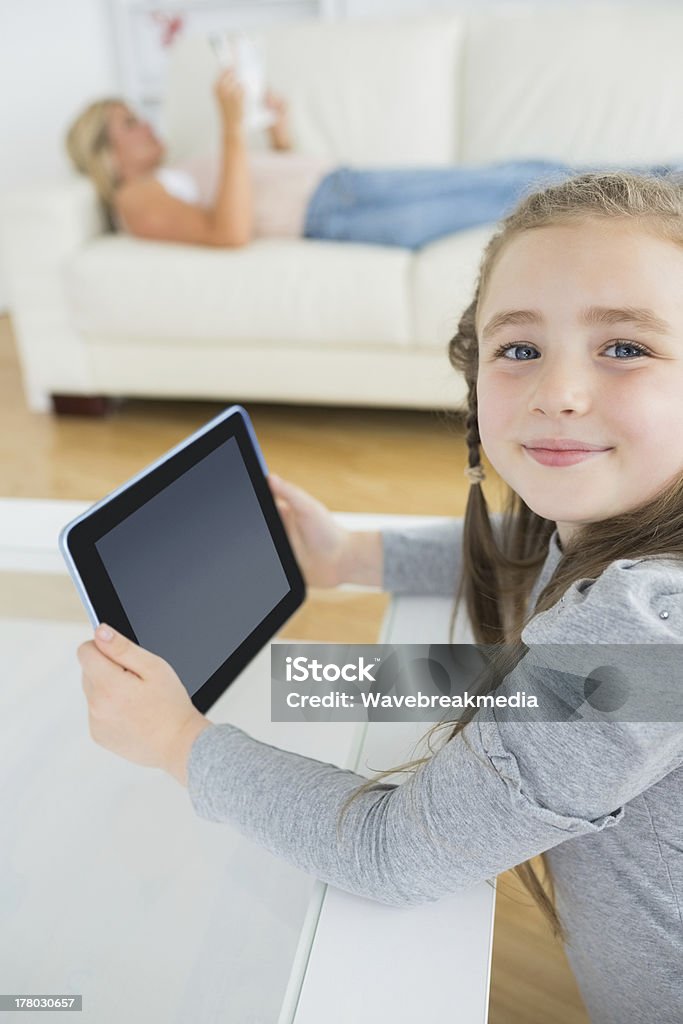 Smiling girl using tablet computer while mother is reading Smiling girl using tablet computer while mother is reading the newspaper on the couch Blond Hair Stock Photo