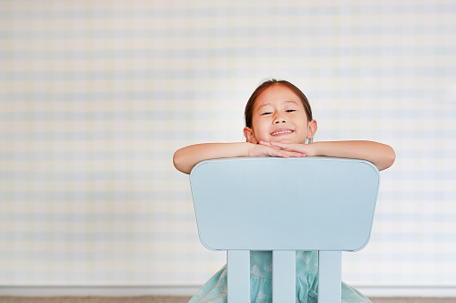 Smiling little Asian child preschool girl in a Kindergarten room poses on plastic baby chair.