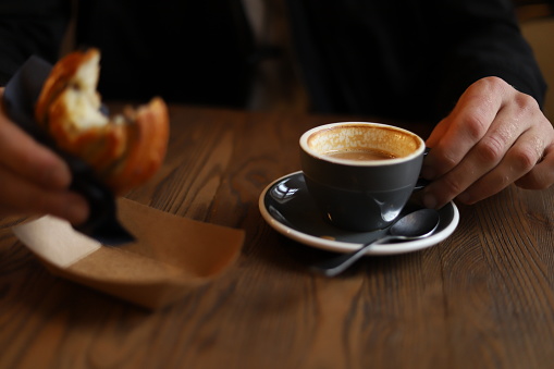 A man's hand holds a round bun and cup of coffee on a wooden table in a cafe. Horizontal indoor photo of snack and fast food concept. Selective focus on hands with blurred background.