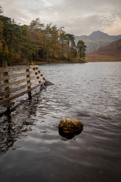 Langdale Pikes from the edge of Blae Tarn. Rock in the foreground and a fence shows the way. Trees line the edge of the lake.