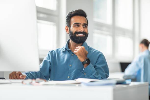 istock Cheerful indian male office worker working on computer and smiling looking aside, sitting at workplace in office 1780363553