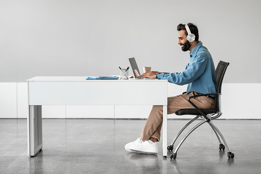 Side view of indian businessman in headphones working on laptop, making video call, sitting at desk, having web conference with customer in office, free space