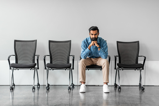 Long time of waiting. Thoughtful serious indian businessman holding hands on chin while sitting on chair against grey wall background, full length, free space
