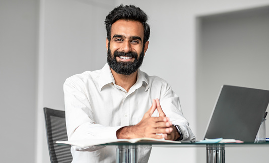 Cheerful confident indian man business consultant working in office, sitting at workdesk in front of laptop computer and smiling at camera, panorama shot