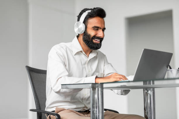 uśmiechnięty indyjski pracownik płci męskiej w słuchawkach bezprzewodowych pracujący na laptopie w biurze, cieszący się pracą online - technology typing male beauty asian and indian ethnicities zdjęcia i obrazy z banku zdjęć