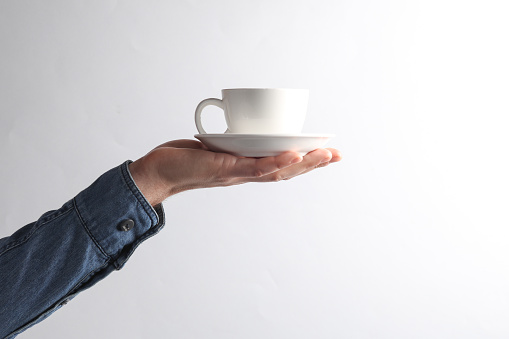 Man's hand in denim shirt holding ceramic cup on a saucer, gray background