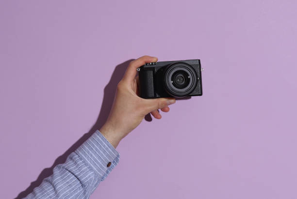 istock Man's hand in shirt holding modern digital camera on purple pastel background with shadow. 1780379909