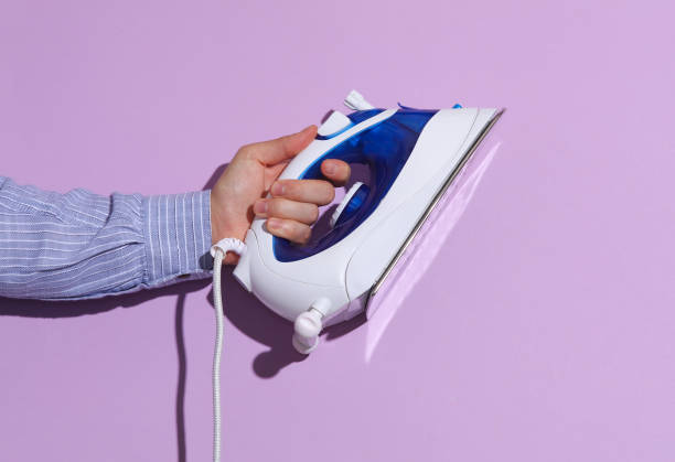 istock Man's hand in shirt holding iron on purple pastel background with shadow. 1780379938