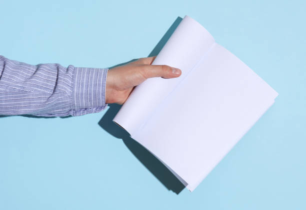 istock Man's hand in shirt holding open white blank magazine mockup on blue background with shadow 1780380514