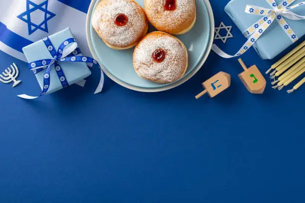 Partake in Hanukkah customs through a top-view picture of sufganiyot, Israeli flag, gift box with bow, candles, and dreidel on a blue background, with space for text or promotional material