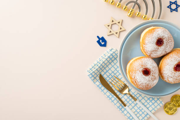 Prepare Hanukkah-themed table arrangement. Top-view image featuring customary Jewish delicacies â sufganiyot, napkin, cutlery, Star of David, menorah, gelt on neutral beige surface with space for text