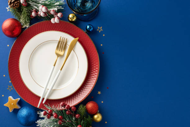 istock Exquisite New Year's Eve restaurant table setting with top view featuring elegant plates, golden flatware, wine glass, ornaments, candle, confetti, frosty fir branches, and mistletoe on blue backdrop 1780388776