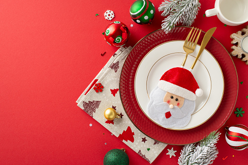Create pleasurable New Year's dinner table setup. Top view of plates, whimsical gold flatware in quirky pocket, napkin, mug, ornaments, snowflake confetti, frosty fir sprigs on red backdrop for promo
