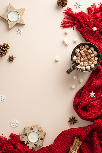 Inviting winter mood: Vertical top view of a cocoa mug with marshmallows, candle, spices, pine cones, snowflakes, and a knitted scarf against a pastel beige background, offering space for text