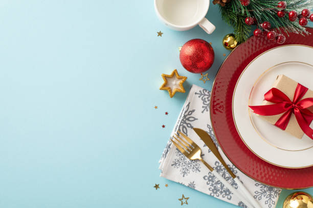 Opt for Christmas-themed dining arrangements. Top view capturing adorned table settings, giftbox, glimmering baubles, winter twig, holly berries on soft blue backdropâideal for text or advert space