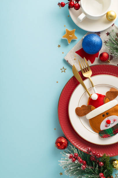 istock Vertical top view delightful holiday party table arrangement with comical cutlery bag, plates, warm mug, colorful baubles, starry candle, frosty fir twigs, berries, pastel blue surface for text or ad 1780388833