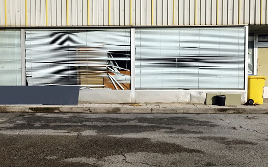 Abandoned store with large windows and damaged venetian blinds, at the roadside. Sidewalk and street in front.