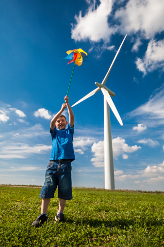 Boy dressed in blue flies a pinwheel on grass near a wind turbine on a hot sunny day with blue sky and white clouds