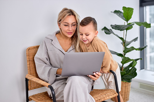 attractive caucasian mother woman and little son using laptop computer together in bright cozy apartment at home. caucasian blonde female work on laptop while child boy stand next to her.