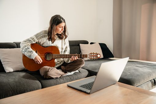 Young woman learning to play guitar at home, sitting on the couch and looking at chords on her laptop. Cozy daytime ambiance in the room.