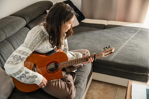 Young woman learning to play guitar at home, sitting on the couch and looking at chords on her laptop. Cozy daytime ambiance in the room.