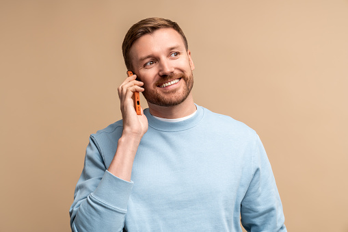 Happy smiling middle aged man talking calling phone on beige background looking aside. Guy enjoying nice pleasant conversation on smartphone. Portrait laughing caucasian male communicating on phone.