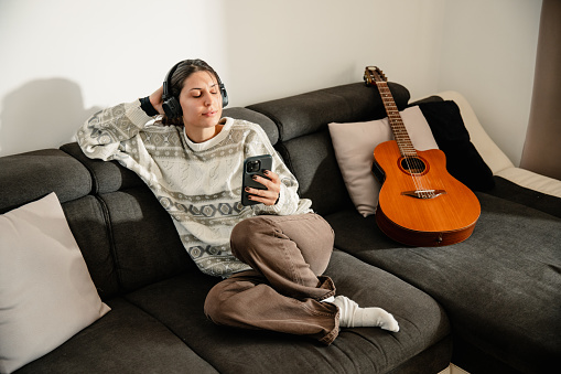 A young and beautiful woman, with headphones, sits on the couch beside an acoustic guitar with a phone in her hand, listening to music, and enjoying a beautiful and pleasant sunny ambiance.
