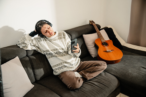 A young and beautiful woman, with headphones, sits on the couch beside an acoustic guitar with a phone in her hand, listening to music, and enjoying a beautiful and pleasant sunny ambiance.