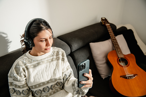 A young and beautiful woman, with headphones, sits on the couch beside an acoustic guitar with a phone in her hand, listening to music, and enjoying a beautiful and pleasant sunny ambiance.