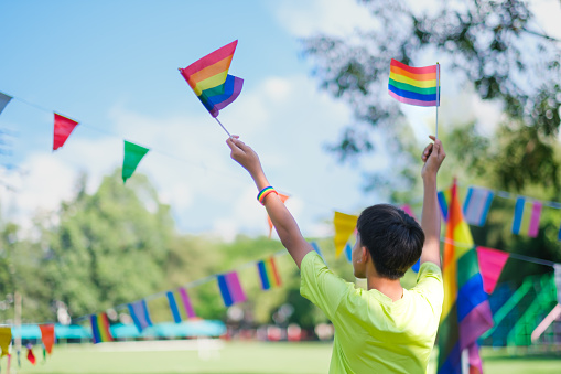 LGBTQ community celebration pride month, young asian kid holding flags facing backwards with nature background. Embracing the lesbian, gay, bisexual, transgender, and queer community.