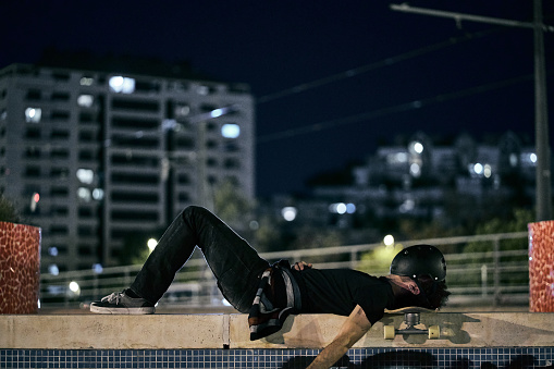 young caucasian skateboarder man lying on a wall in the quiet of the night in the city with the skateboard under his head and the helmet on his face - Lfestyle concept