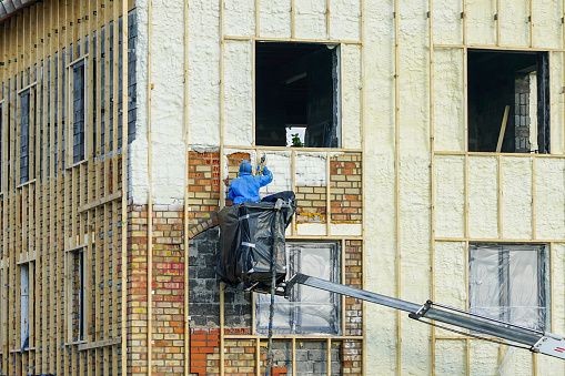 Technician spraying thermal insulation foam layer on the exterior wall of a residential house using plural component gun for polyurethane foam