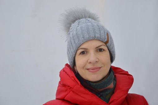 Portrait of a young Caucasian woman in a red jacket with a hood and a gray winter knitted hat on a light background.